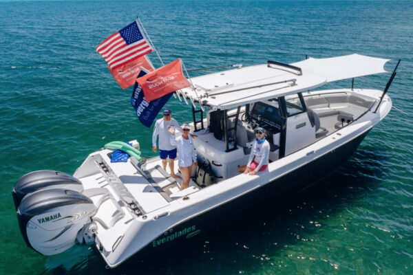 DJI_0687_8614 An Everglades boat floating on clear water with three people on board waving, multiple flags flying from the back, and two Yamaha engines at the stern