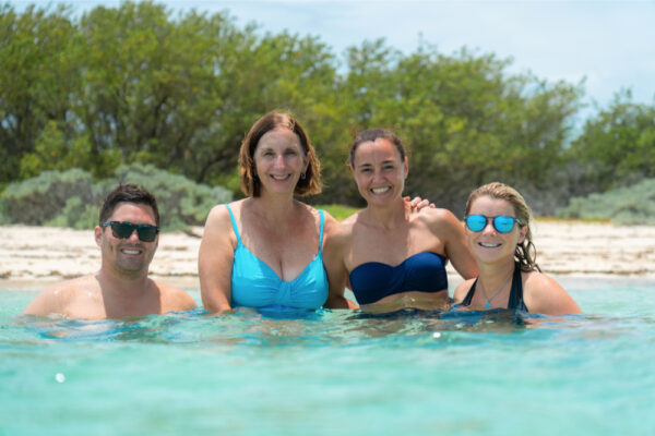 JJS00169_3973 Four people smiling and standing in shallow turquoise water near a sandy shoreline with green shrubs in the background