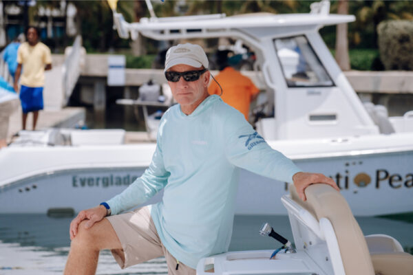 JJS06940_1837 A man wearing sunglasses and a light blue shirt leaning on an Everglades boat at a marina with another Everglades boat in the background