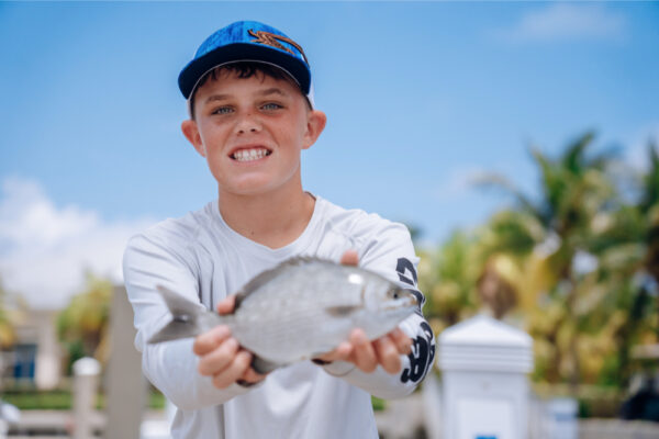 JJS06947_9821 A young boy smiling and holding a small fish in his hands, wearing a long-sleeve shirt and a blue cap, with a sunny marina and palm trees in the background