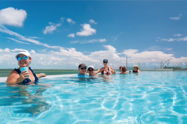 JJS07034_1371 A group of people relaxing in a pool on a sunny day with clear blue skies and scattered clouds wearing hats and sunglasses while enjoying drinks