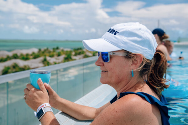 JJS07039_4391 A woman wearing an Everglades hat and sunglasses relaxing in a pool while holding a drink with a scenic view of palm trees and the ocean in the background under a partly cloudy sky