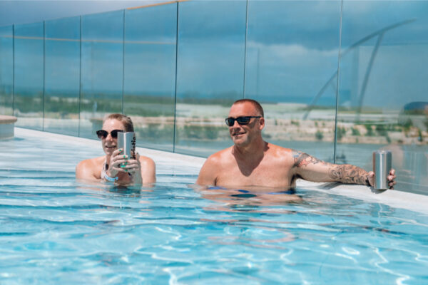 JJS07044_2525 Two people relaxing in a pool holding stainless steel tumblers and wearing sunglasses with a glass fence and a landscape visible in the background