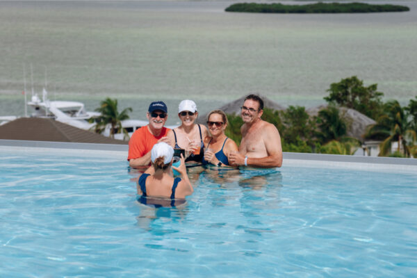 JJS07072_4881 A group of five people standing in an infinity pool overlooking a scenic view of the ocean and trees, smiling and enjoying drinks together