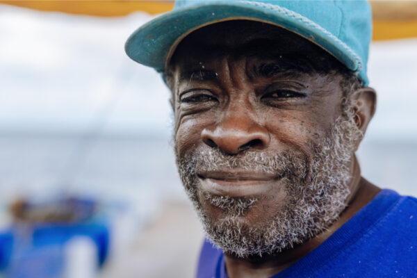JJS07322_859 A close-up of a man with a beard wearing a blue hat and shirt, smiling slightly, with a blurred background of blue and yellow tones