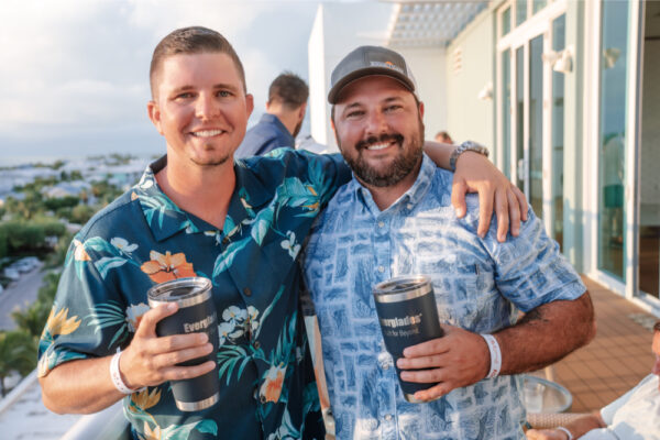 JJS07391_1377 Two men smiling and standing together on a deck, each holding an Everglades-branded tumbler, wearing casual floral shirts during a gathering