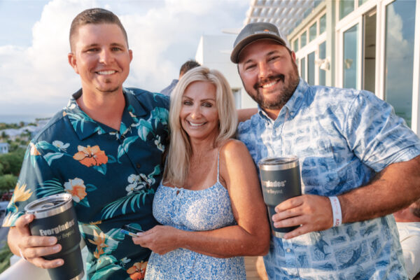 JJS07393_4036 Two men and a woman smiling together on a deck, with the men holding Everglades-branded tumblers, wearing casual floral shirts during a gathering