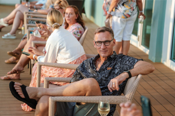 JJS07394_4061 A man sitting in a chair on an outdoor deck wearing glasses and a patterned shirt with other people seated and talking in the background