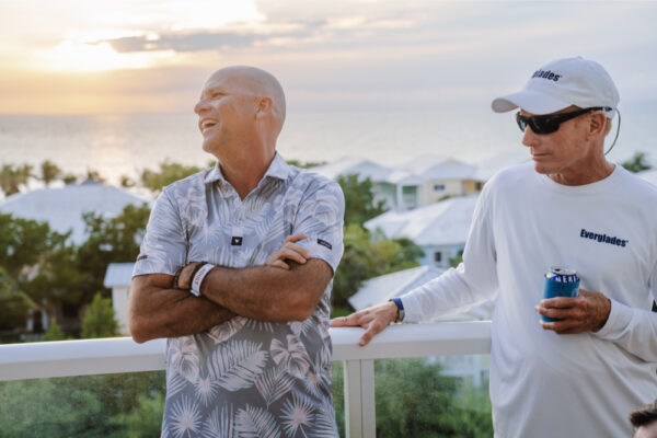 JJS07414_9328 Two men standing on a balcony at sunset with one man smiling and the other holding a drink while wearing an Everglades shirt and sunglasses with a scenic view of rooftops and trees in the background