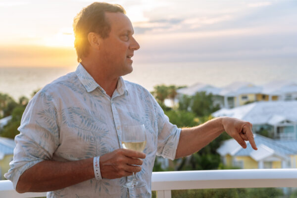 JJS07435_274 A man holding a glass of wine while standing on a balcony at sunset pointing and talking with a scenic view of rooftops and the ocean in the background
