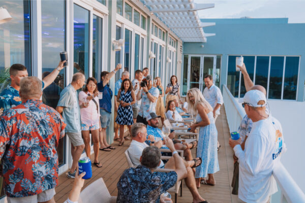 JJS07522_8794 A group of people gathered on a large outdoor deck raising drinks and toasting with some seated and others standing during a casual event at a building with large windows