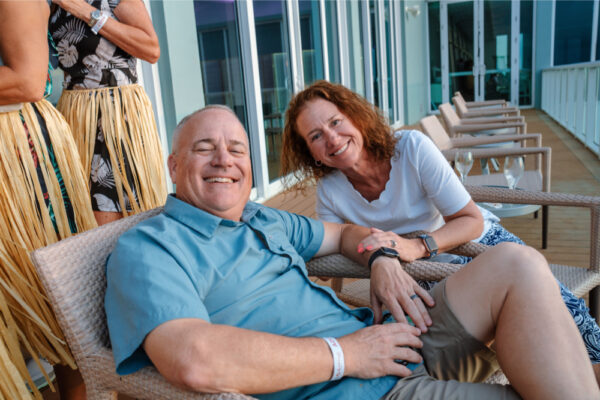 JJS07551_3337 A man and woman sitting and smiling on an outdoor deck during a casual gathering with other people standing in the background wearing grass skirts