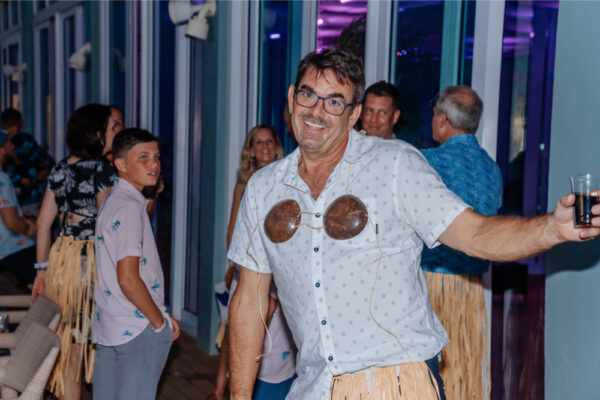 JJS07562_1783 A man smiling and posing for the camera at a party wearing a coconut bra and grass skirt with other people in the background