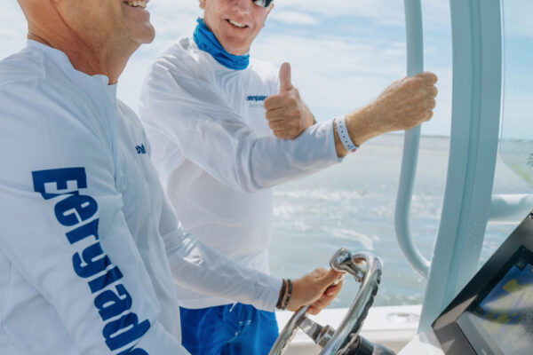 JJS09693_9723 Two men wearing sunglasses and white shirts steering a boat, with one man giving a thumbs up gesture
