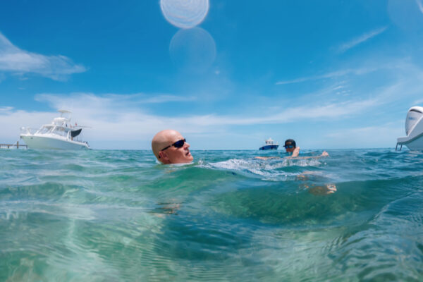 UW8096_5425 Two people swimming in clear turquoise water with Everglades boats in the background under a bright blue sky