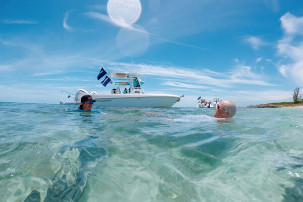 UW8099_9378 Two people swimming in clear turquoise water with an Everglades boat floating nearby under a bright blue sky