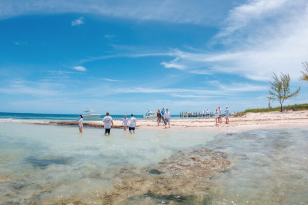 UW8120_9820 A group of people wading in shallow water near a sandy beach with boats docked in the distance under a bright blue sky